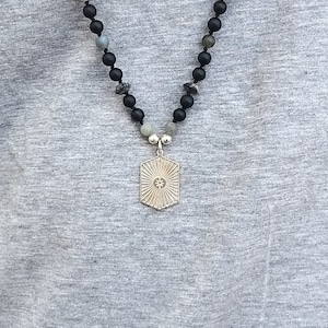 May include: A silver pendant necklace with a geometric starburst design hangs from a black beaded chain. The beads are a mix of black and gray.