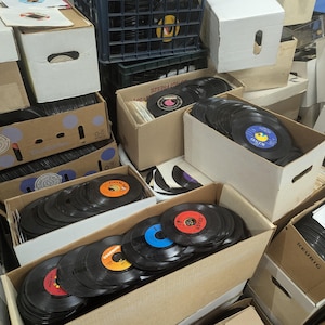 Könnte beinhalten: Eine Sammlung von Schallplatten in Pappkartons. Die Schallplatten haben farbenfrohe Etiketten und sind in verschiedenen Kartons gestapelt. Die Kartons sind unterschiedlich groß und geformt und bilden eine unordentliche, aber organisierte Präsentation.