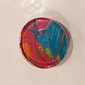 May include: A round, colorful coaster with a swirling pattern of red, blue, green, yellow, and white paint. The coaster has a glossy finish.
