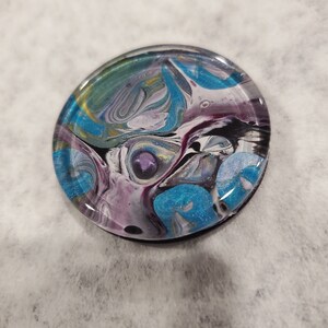 May include: A round, clear resin coaster with a swirling abstract design in shades of blue, purple, white, black, and gold.