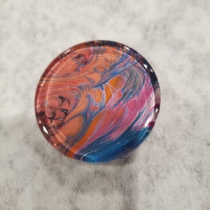 May include: A round, clear glass coaster with a colorful abstract design. The design features swirls of orange, pink, blue, and black. The coaster is set against a white, textured background.