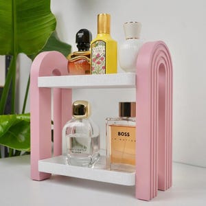 May include: A two-tiered pink arched shelf with white speckled surfaces holding five glass perfume bottles. The top shelf features a black, yellow, and white bottle, while the bottom shelf displays a clear bottle and a rectangular amber bottle.