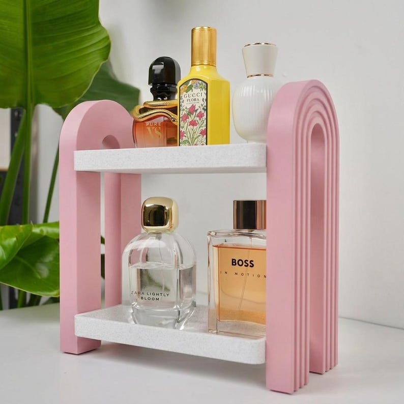 May include: A two-tiered pink arched shelf with white speckled surfaces holding five glass perfume bottles. The top shelf features a black, yellow, and white bottle, while the bottom shelf displays a clear bottle and a rectangular amber bottle.