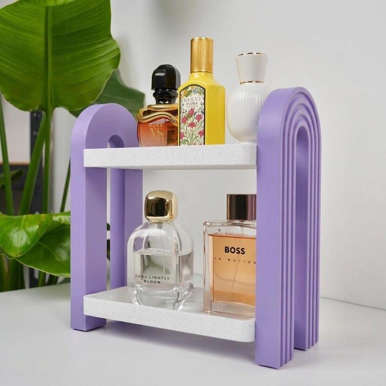 May include: A two-tiered storage shelf with purple arched side supports and white speckled shelves. The top shelf holds a black and gold perfume bottle, a yellow Gucci Flora bottle, and a white bottle. The bottom shelf holds two glass perfume bottles.