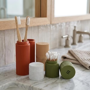 May include: A collection of cylindrical bathroom organisers with a vertical ribbed texture. The set includes a tall terracotta holder with two bamboo toothbrushes, a tan container, a white lidded jar, and a green jar filled with cotton swabs.