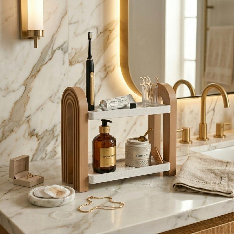 May include: A two-tier bathroom organizer with light wood arched side supports and white speckled shelves. The top shelf holds a black electric toothbrush and a tube of toothpaste. The bottom shelf holds a brown glass soap bottle and a white jar.