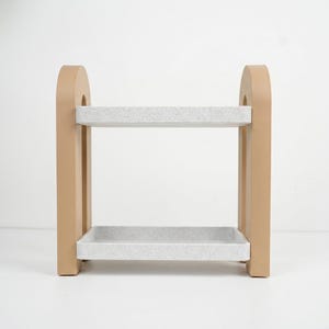 May include: A two-tiered storage shelf featuring two rectangular, light gray speckled trays supported by two solid, smooth, tan-colored arched side panels. The structure is open on all sides, providing a clear view of the two horizontal levels.