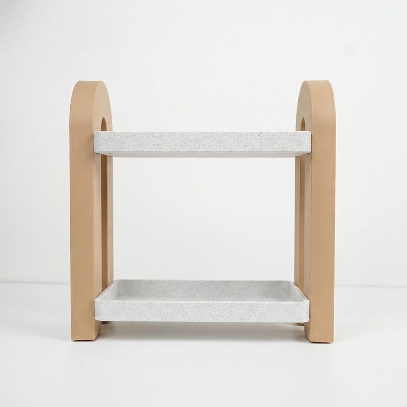 May include: A two-tiered storage shelf featuring two rectangular, light gray speckled trays supported by two solid, smooth, tan-colored arched side panels. The structure is open on all sides, providing a clear view of the two horizontal levels.