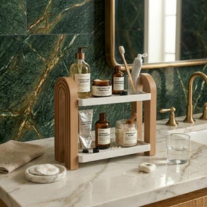 May include: A two-tiered bathroom organizer with light brown arched side supports and white shelves. It holds various skincare products including hand wash, hand balm, bath essence, Epsom salt, a tube of hand cream, and a toothbrush in a glass.