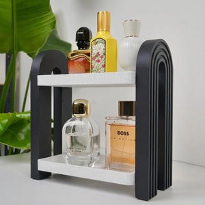 May include: A two-tier shelf featuring black arched side supports with ribbed detailing and two white speckled rectangular shelves. The unit holds five glass perfume bottles of varying shapes and sizes, including brands like Gucci, Zara, and Boss.