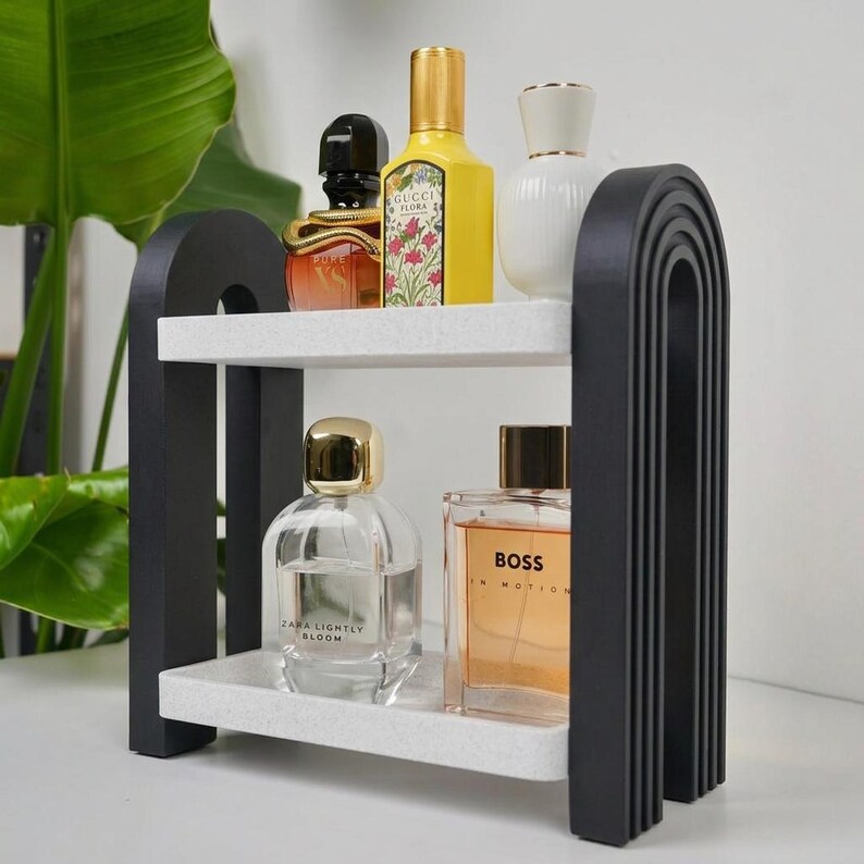 May include: A two-tier shelf featuring black arched side supports with ribbed detailing and two white speckled rectangular shelves. The unit holds five glass perfume bottles of varying shapes and sizes, including brands like Gucci, Zara, and Boss.