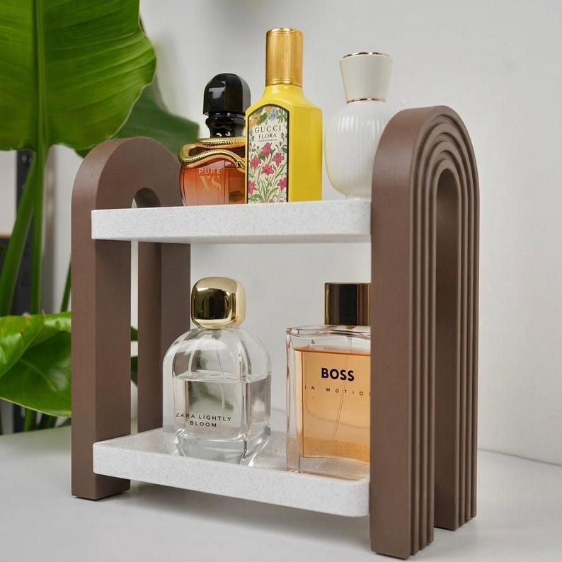 May include: A two-tiered storage shelf featuring brown arched side supports with ribbed detailing and white speckled rectangular shelves. The top shelf holds a black and gold perfume bottle, a yellow Gucci Flora bottle, and a white bottle. The bottom shelf holds a clear Zara Lightly Bloom bottle and a rectangular Boss In Motion bottle.