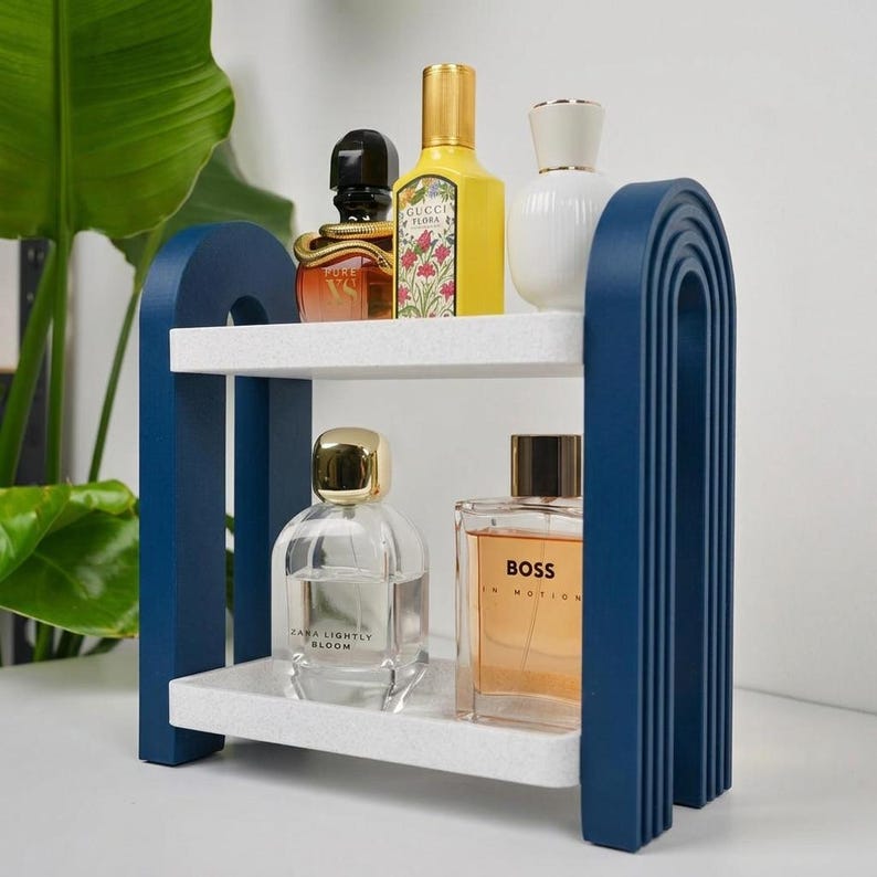 May include: A two-tiered shelf with white speckled shelves and dark blue arched side supports. The top shelf holds a black bottle, a yellow Gucci Flora bottle, and a white bottle. The bottom shelf holds a clear Zara bottle and a Boss bottle.