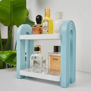 May include: A two-tiered light blue arched shelf with white speckled surfaces holding five glass perfume bottles. The top shelf features a black, a yellow, and a white bottle, while the bottom shelf holds two clear glass bottles with gold caps.