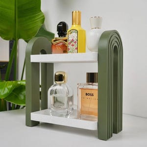 May include: A two-tiered shelf with olive green arched side supports and speckled white rectangular shelves. The top shelf holds a black and gold perfume bottle, a yellow Gucci Flora bottle, and a white bottle. The bottom shelf holds two glass perfume bottles.