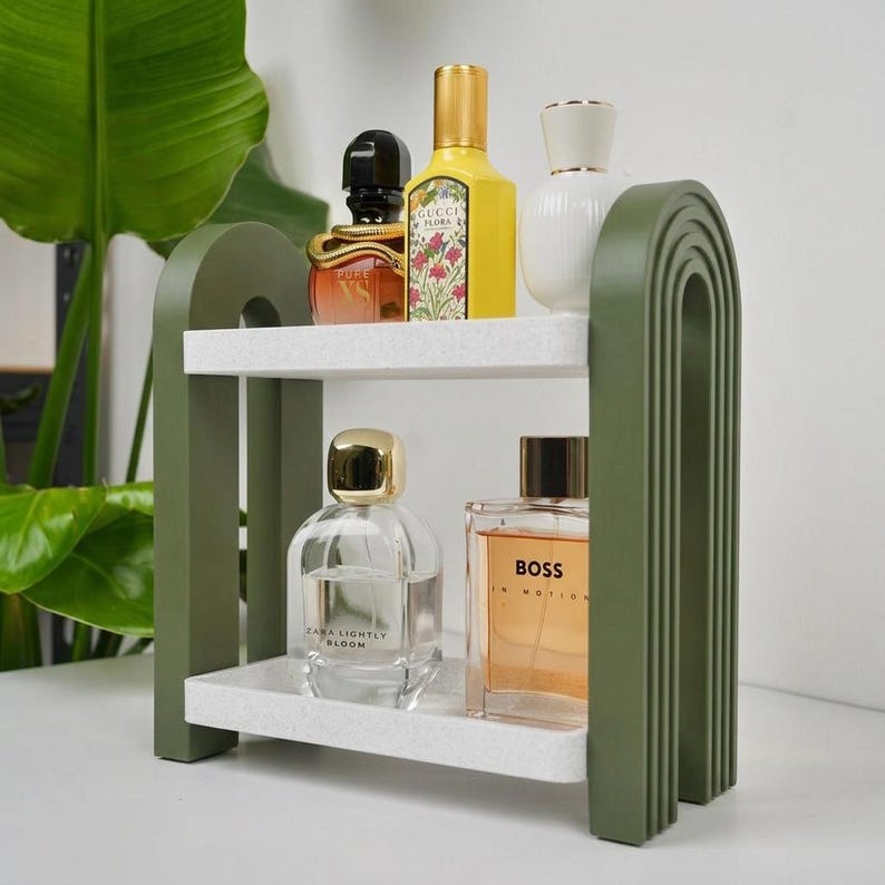 May include: A two-tiered shelf with olive green arched side supports and speckled white rectangular shelves. The top shelf holds a black and gold perfume bottle, a yellow Gucci Flora bottle, and a white bottle. The bottom shelf holds two glass perfume bottles.
