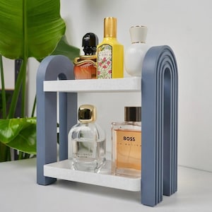 May include: A two-tier storage shelf with a blue arched frame and speckled white shelves. The top shelf holds a black, a yellow Gucci Flora, and a white perfume bottle. The bottom shelf holds a clear Zara Lightly Bloom and a Boss In Motion bottle.