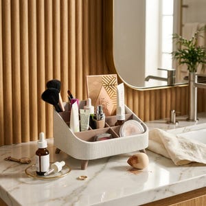 Peut inclure: Un organisateur de cosmétiques crème à compartiments multiples, doté d'un extérieur nervuré et de quatre petits pieds marron. Il contient divers pinceaux de maquillage, tubes, flacons et une palette marquée 'Lust Nude' sur un comptoir en marbre.
