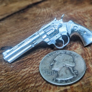 May include: A silver-colored revolver replica, approximately the size of a US quarter, on a wooden surface.