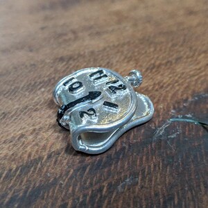 May include: A silver-colored, melting clock pendant with black clock hands and numerals. The clock face shows the numbers 10, 11, 12, 1, and 2. The pendant has a loop at the top for a chain.