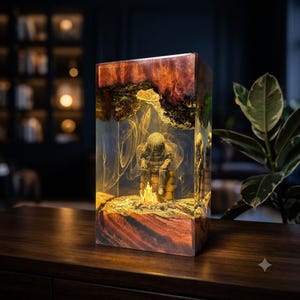 Könnte beinhalten: Eine rechteckige Lampe aus dunklem Holz und klarem Harz mit einer Innenszene einer Figur, die an einem Lagerfeuer sitzt. Das Harz enthält wirbelnde goldene Lichtmuster und das Holz weist eine natürliche, strukturierte Maserung auf.