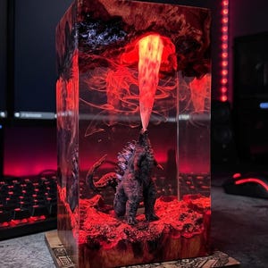 May include: A rectangular resin lamp featuring a Godzilla figure standing on a textured red base. The creature is positioned beneath a dark, cavernous top section, emitting a bright, glowing red beam of light upward into the resin structure.