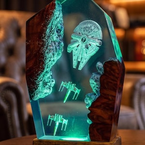 Könnte beinhalten: Eine vertikale, fünfeckige Harzlampe mit eingebetteten hölzernen Felsformationen und Miniatur-Raumschiffen, darunter ein Millennium Falke und zwei X-Wing-Jäger. Das Objekt wird grün beleuchtet und steht auf einem rechteckigen Holzsockel.