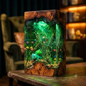 May include: A rectangular wooden lamp featuring a clear resin centre filled with a miniature forest scene. Inside the resin, a green creature with a bulbous plant on its back sits among small colourful mushrooms, vines, and moss, illuminated by a bright green light.