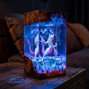 Pode incluir: Uma luminária retangular com uma figura branca de Mewtwo encapsulada em resina transparente entre duas peças de madeira escura e texturizada. O interior é iluminado com uma luz azul vibrante, criando um efeito de fumaça ao redor da figura.