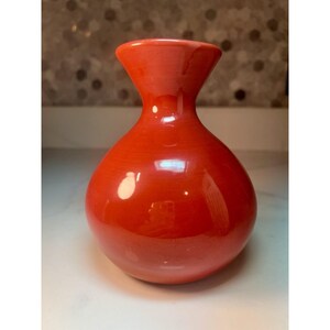May include: A bright orange ceramic vase with a smooth, glossy finish. The vase features a wide, rounded base that tapers into a narrow neck, ending in a flared, circular opening at the top. The surface reflects light, showing a polished texture.