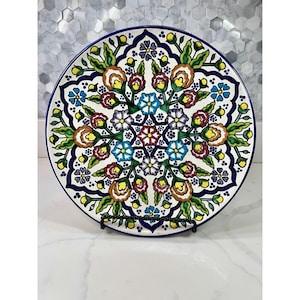 May include: A round ceramic decorative plate featuring a symmetrical hand-painted floral pattern. The design includes intricate motifs in shades of cobalt blue, deep red, bright yellow, and green against a white background, framed by a dark blue border.