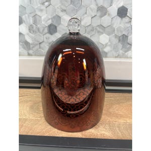 May include: A dome-shaped glass cloche with a deep amber, reflective finish. The surface features a repeating, subtle geometric quatrefoil pattern etched into the glass. A small, clear spherical knob sits at the very top of the rounded dome.