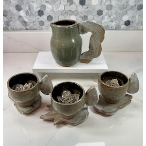 May include: A handmade ceramic pitcher and three matching cups. The pitcher is olive green with a leaf-shaped handle. The cups are brown and gray, each with a leaf-shaped accent and a clear crystal inside. The set is displayed on a white surface.