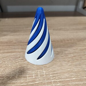 May include: A conical object featuring a blue core with a spiraling white outer shell. The white exterior has diagonal cutouts that reveal the solid blue interior, creating a distinct twisted pattern that tapers to a rounded point at the top.