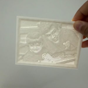 May include: A rectangular white 3D printed lithophane plaque featuring a relief image of two individuals. The surface displays a detailed monochromatic portrait of a child and an adult, with visible textures and depth created by varying plastic thickness.