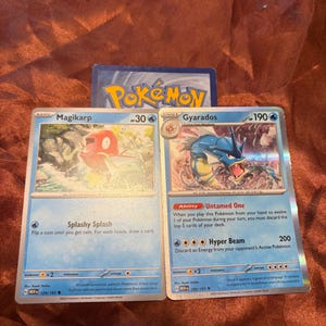 May include: Two Pokémon trading cards featuring Magikarp and Gyarados. The Magikarp card shows a red fish in water with the text Splashy Splash. The Gyarados card depicts a blue sea serpent with the ability Untamed One and the attack Hyper Beam.