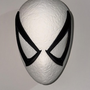 May include: A white mask with black accents, featuring large, stylized eye openings with a mesh texture. The mask has a textured surface and a smooth, rounded shape, suggesting a superhero or cosplay theme.