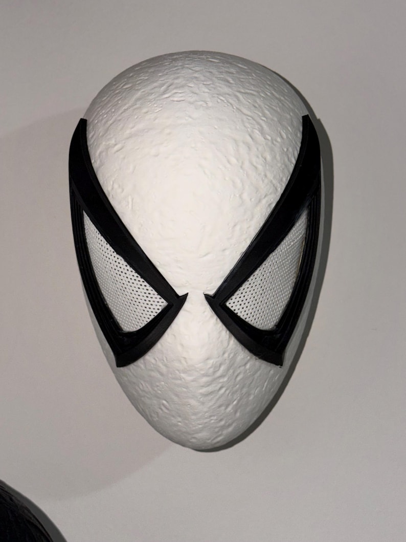 May include: A white mask with black accents, featuring large, stylized eye openings with a mesh texture. The mask has a textured surface and a smooth, rounded shape, suggesting a superhero or cosplay theme.