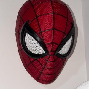 May include: A red and black Spider-Man mask with white mesh eye coverings. The mask has a textured surface with a web pattern. The eye frames are black, and the mask is mounted on a white wall.