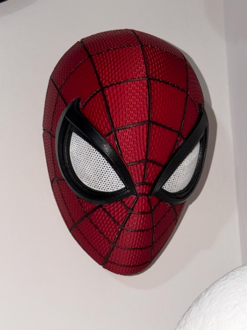 May include: A red and black Spider-Man mask with white mesh eye coverings. The mask has a textured surface with a web pattern. The eye frames are black, and the mask is mounted on a white wall.