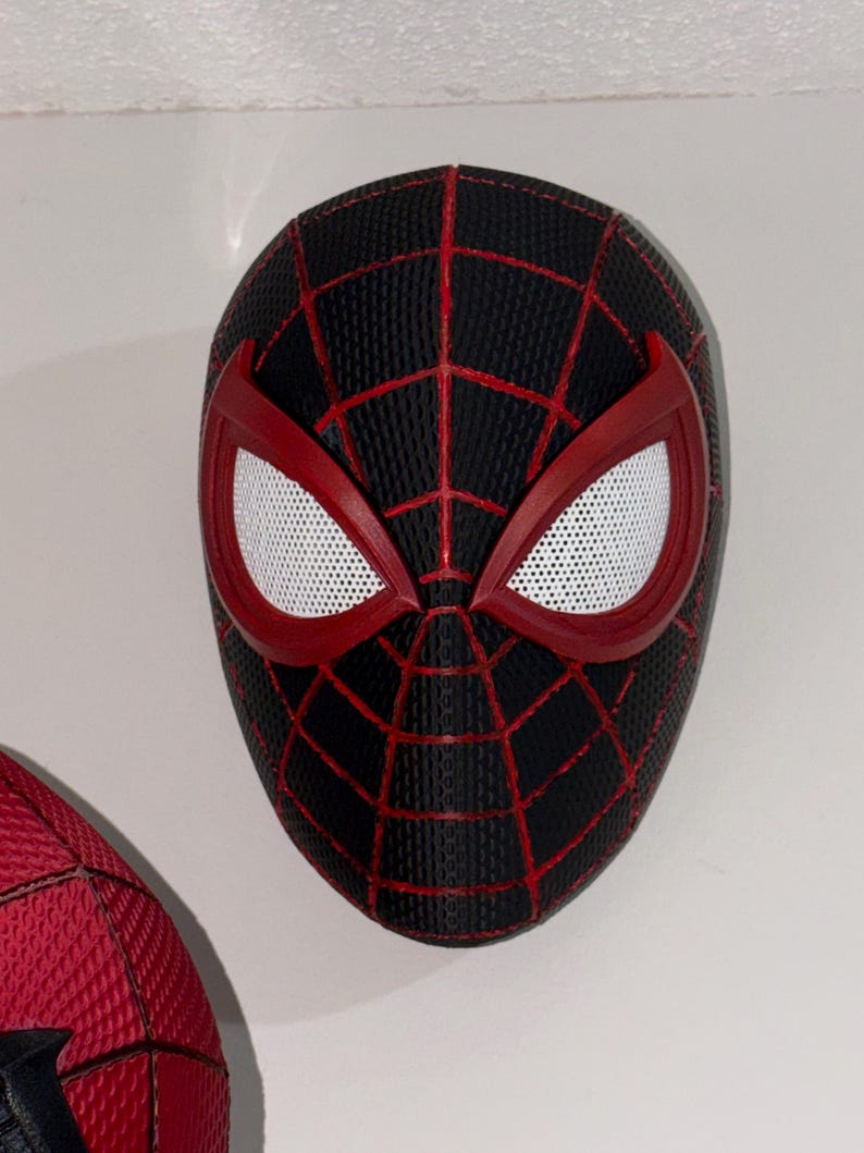 May include: A black and red Spider-Man mask with white mesh eye coverings. The mask has a textured surface and a web-like pattern in red. The eye frames are red.