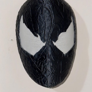 May include: A black mask with white eye openings. The mask has a textured surface and a design reminiscent of a comic book character. The eye openings are white with small perforations.