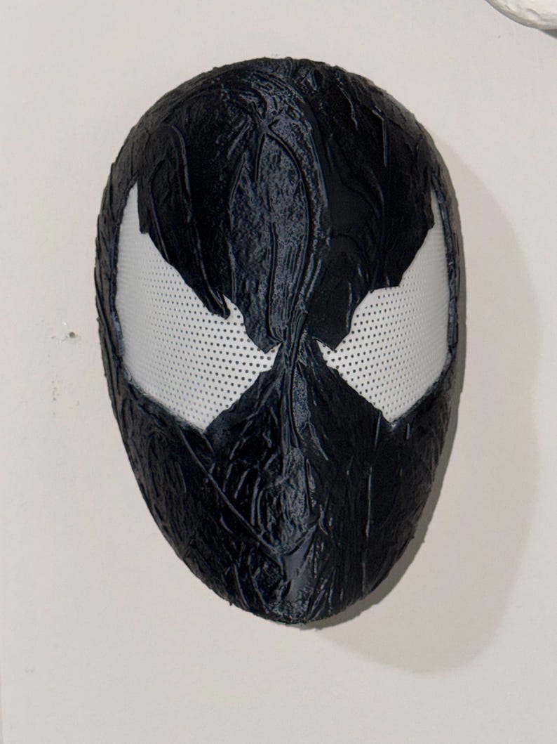 May include: A black mask with white eye openings. The mask has a textured surface and a design reminiscent of a comic book character. The eye openings are white with small perforations.