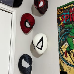 May include: Five Spider-Man masks in various designs are mounted on a white wall. The masks include red, black, and white color schemes, with web patterns and eye details. A vintage comic book poster is visible on the right.