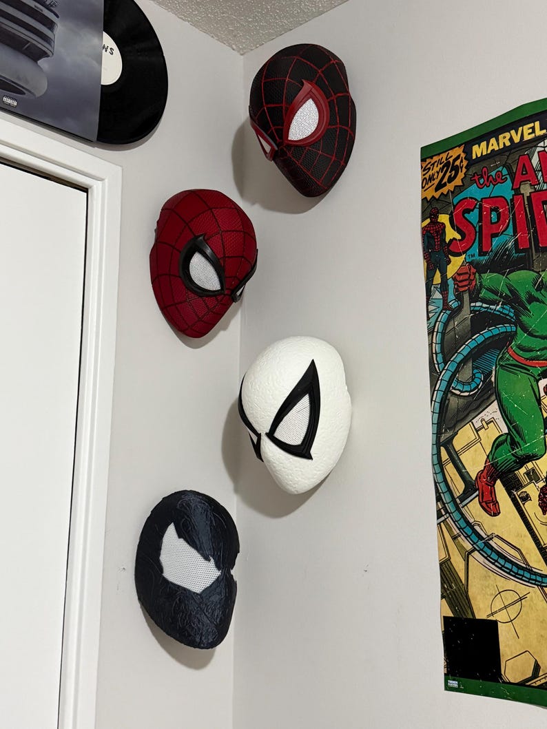May include: Five Spider-Man masks in various designs are mounted on a white wall. The masks include red, black, and white color schemes, with web patterns and eye details. A vintage comic book poster is visible on the right.
