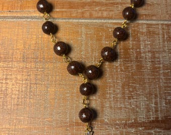 Peut inclure: Un chapelet composé de perles sphériques marron foncé mouchetées, reliées par des maillons en métal doré. Le chapelet se termine par un pendentif en forme de croix dorée, orné de petits cristaux transparents et d'une perle blanche centrale.
