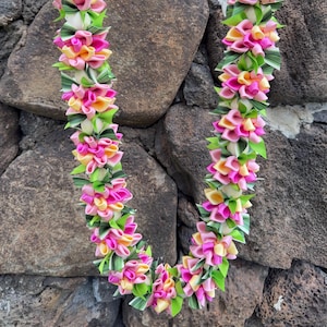 May include: A decorative lei featuring a repeating pattern of folded ribbon flowers in shades of bright pink, soft yellow, and light green, interspersed with green striped ribbon leaves and white spherical beads, suspended by a lime green ribbon.