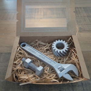 May include: A brown cardboard gift box with a clear window containing three silver-coloured items nestled in shredded paper: an 8-inch adjustable wrench with the text "DROP FORGED" embossed on the handle, a metal bolt with a nut, and a gear.
