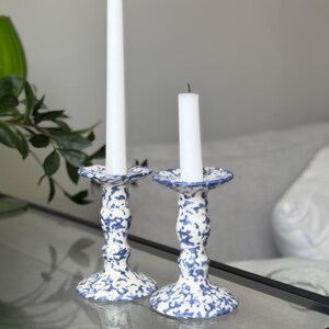 Può includere: Due portacandele in ceramica con un motivo maculato blu e bianco. Ogni portacandele presenta una base svasata, uno stelo sottile con un dettaglio centrale bombato e un bordo superiore largo. Una candela bianca è inserita in ogni supporto.