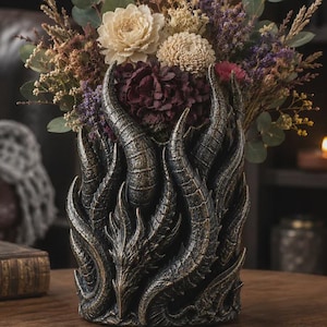 Könnte beinhalten: Eine zylindrische Vase mit dunkler, verwitterter Metalloberfläche, verziert mit erhabenen drachenartigen Schuppen und geschwungenen Hornmotiven. Das Gefäß ist mit einem Strauß aus getrockneten Blumen in Creme-, Lila- und Grüntönen gefüllt.